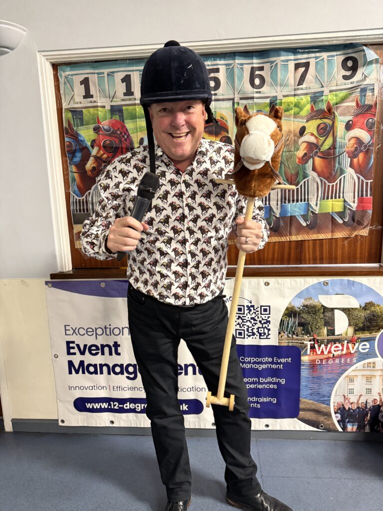 Sussex Race Nights host presenting with a microphone and toy horse in front of a tote board banner